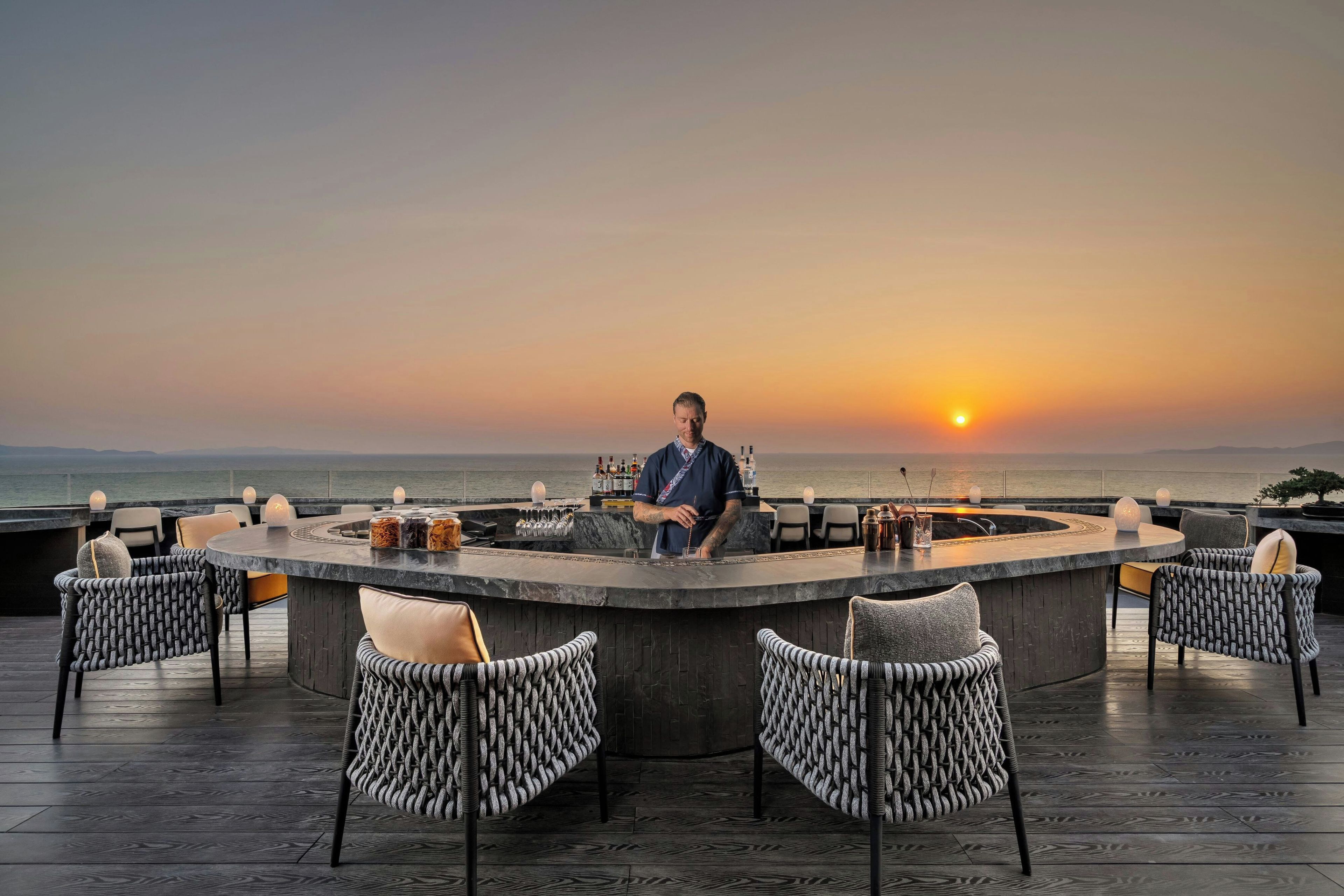 A bar area with a view of the sunset over the ocean, equipped with bar stools and a bartender.