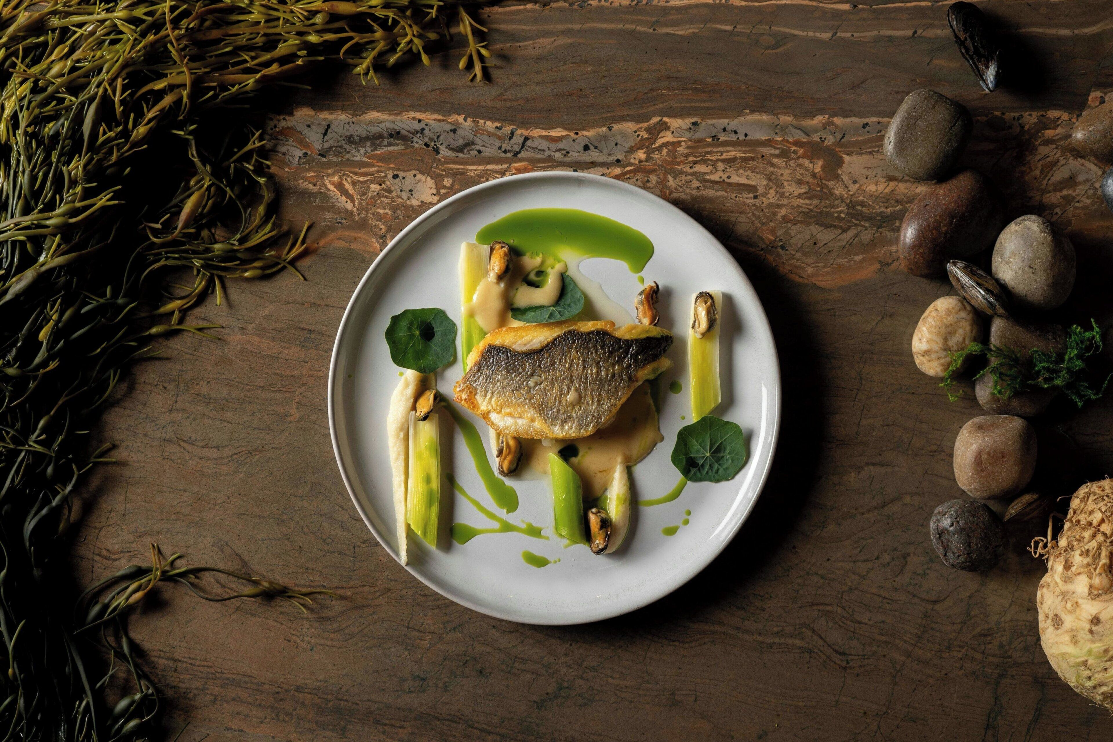 Une assiette de nourriture comprenant un poisson, des asperges et une sauce verte, posée sur une table en bois.