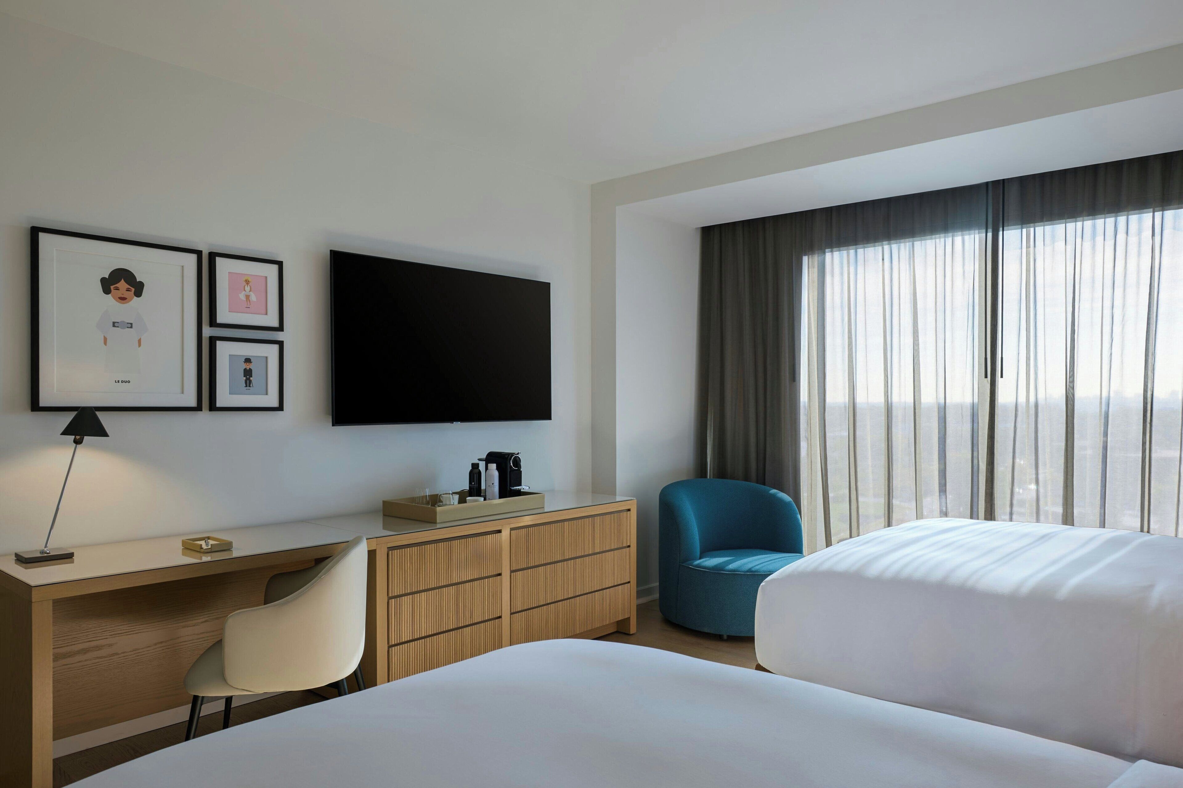Habitación de hotel con cama, escritorio con silla, televisor, sillón azul y una ventana grande con cortinas.