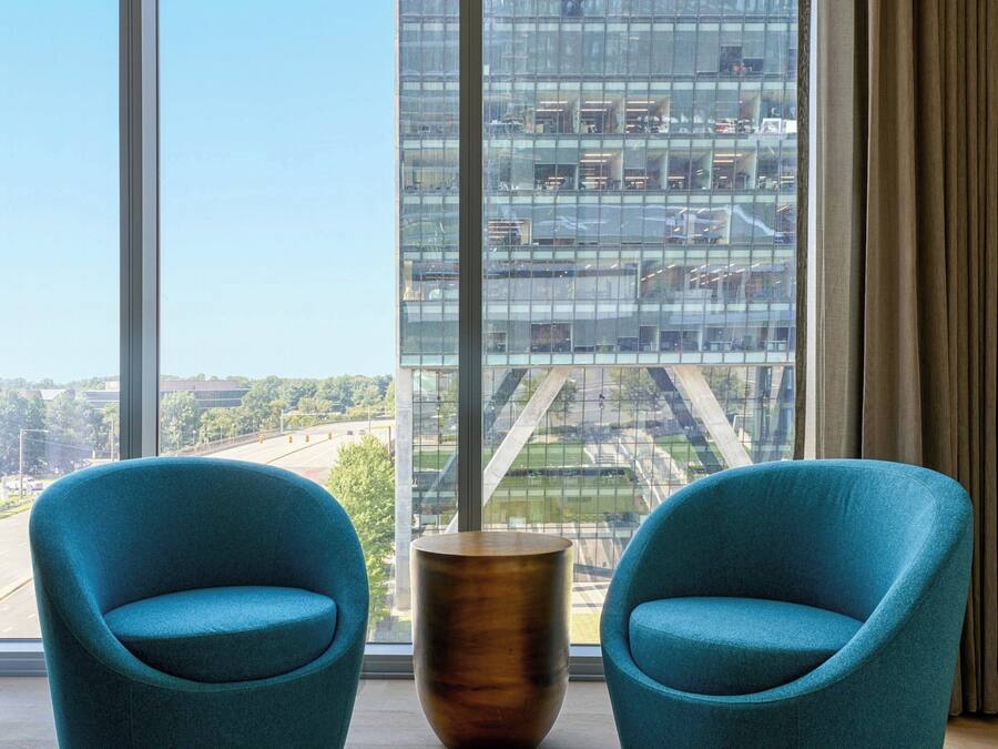 A room with two teal chairs, a wooden side table, and a large window offering a view of a modern building and a bridge.
