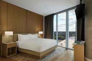 A hotel room with a large bed, two bedside tables with lamps, a view of the city through large windows, and a small wooden side table with books.
