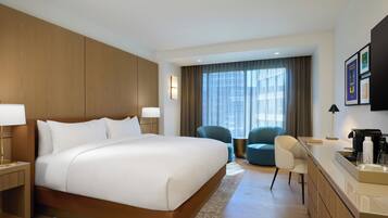 Habitación de hotel con una cama grande, un escritorio con cafetera, una silla y vistas de la ciudad a través de amplios ventanales.