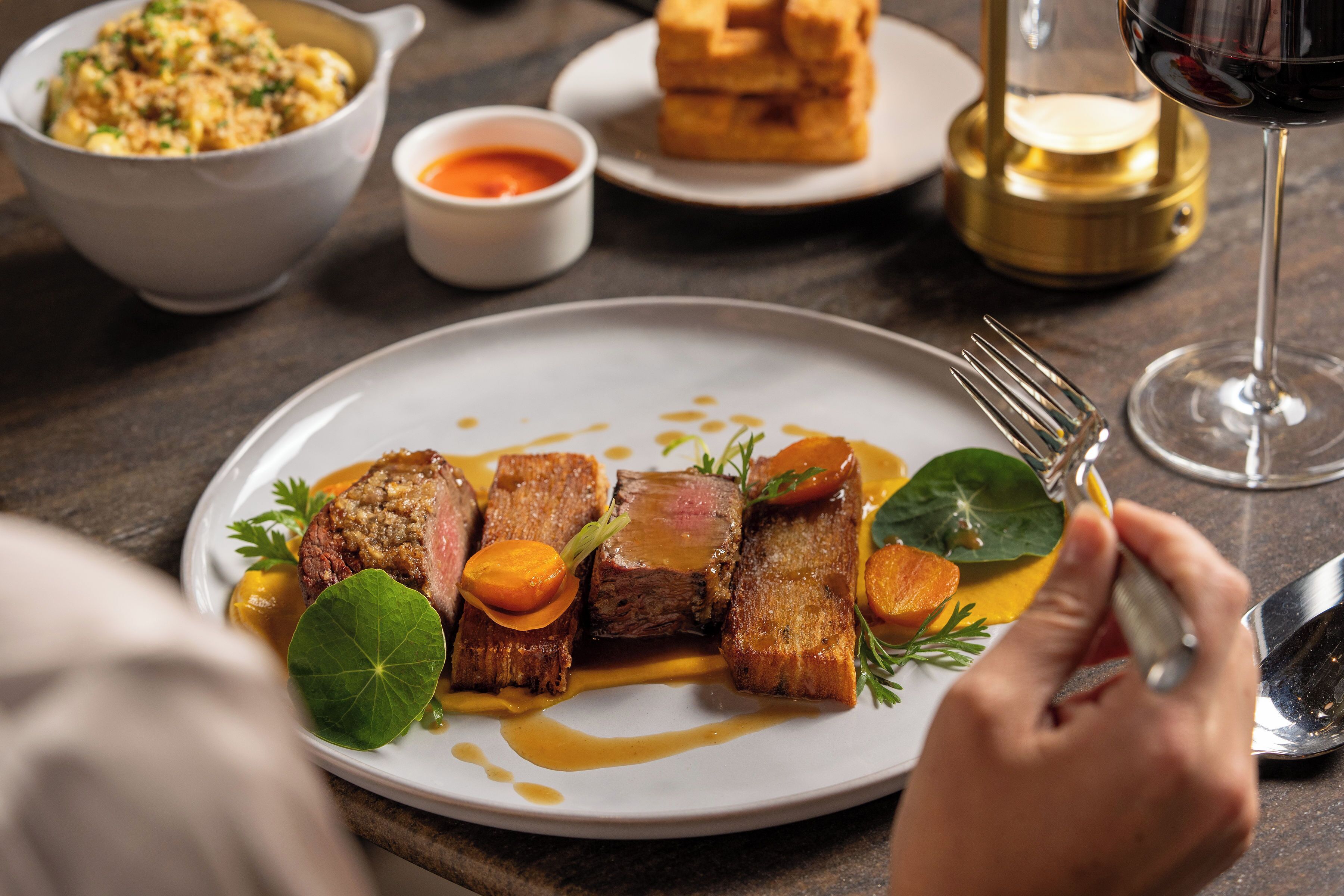 Une assiette de nourriture avec une fourchette et un couteau, un verre de vin rouge et un bol de soupe.