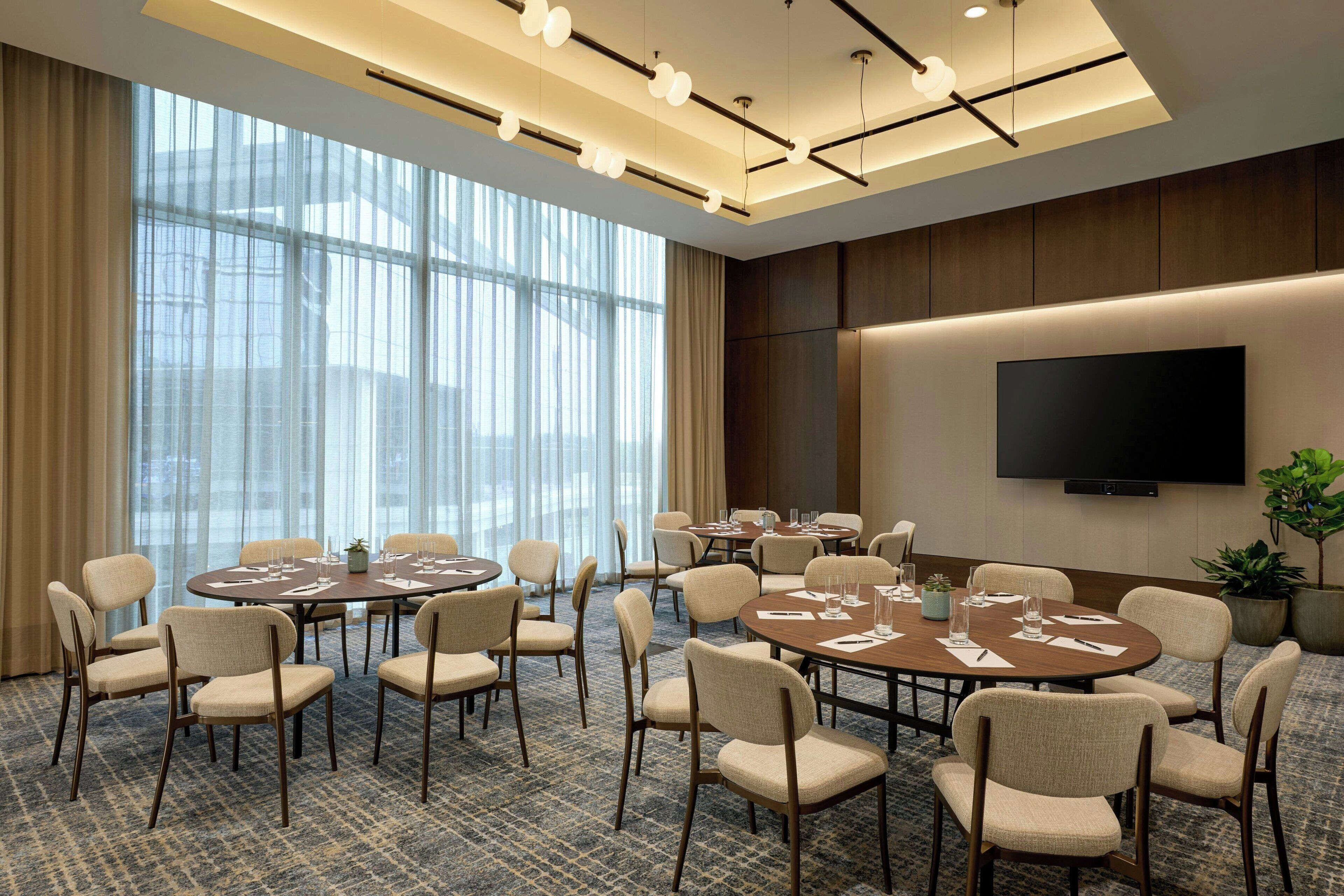 A conference room with multiple tables and chairs, large windows, and a flat-screen TV on the wall.