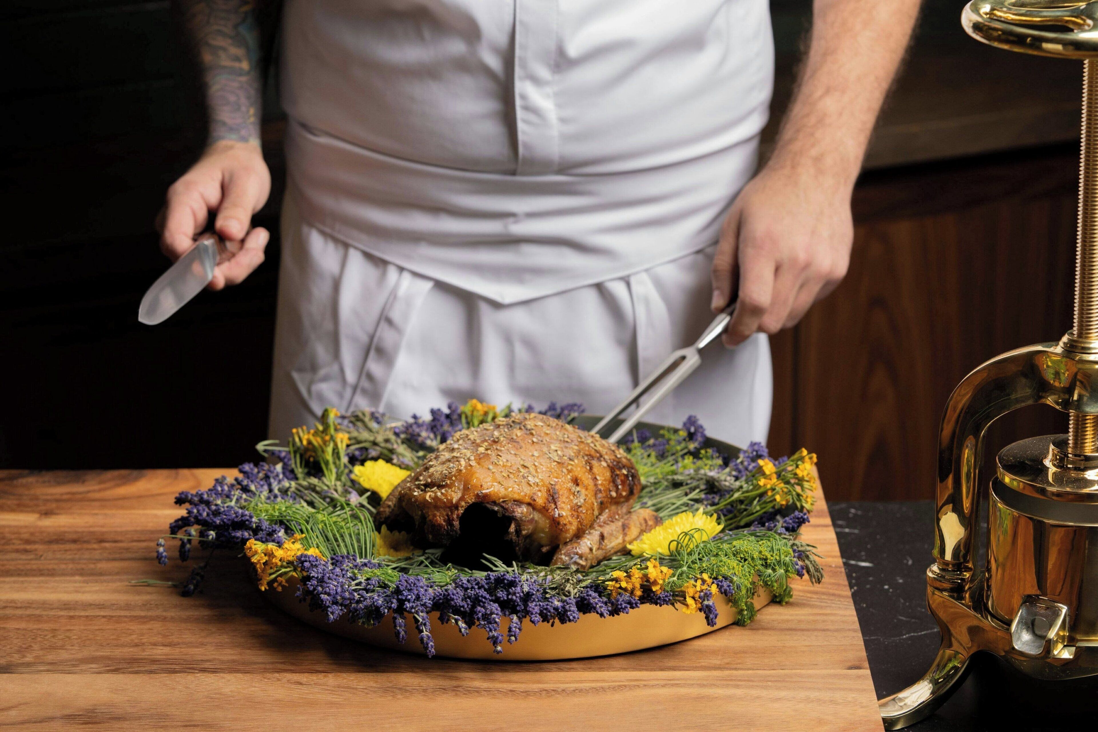 A roasted turkey on a wooden platter surrounded by herbs and flowers.