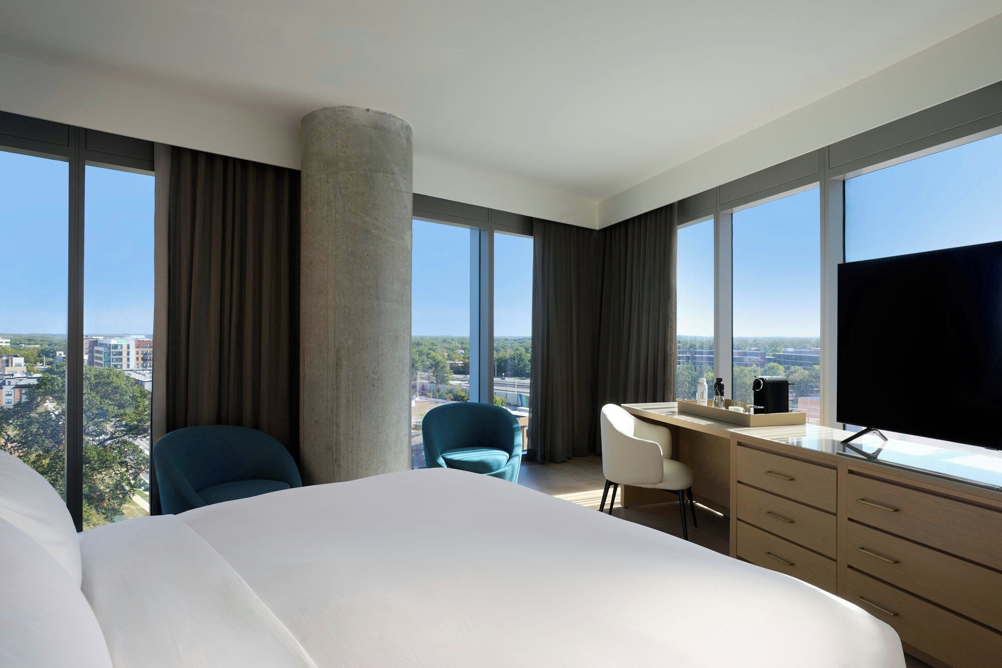 A hotel room with a large bed, a desk, a chair, a TV, and a view of the city.