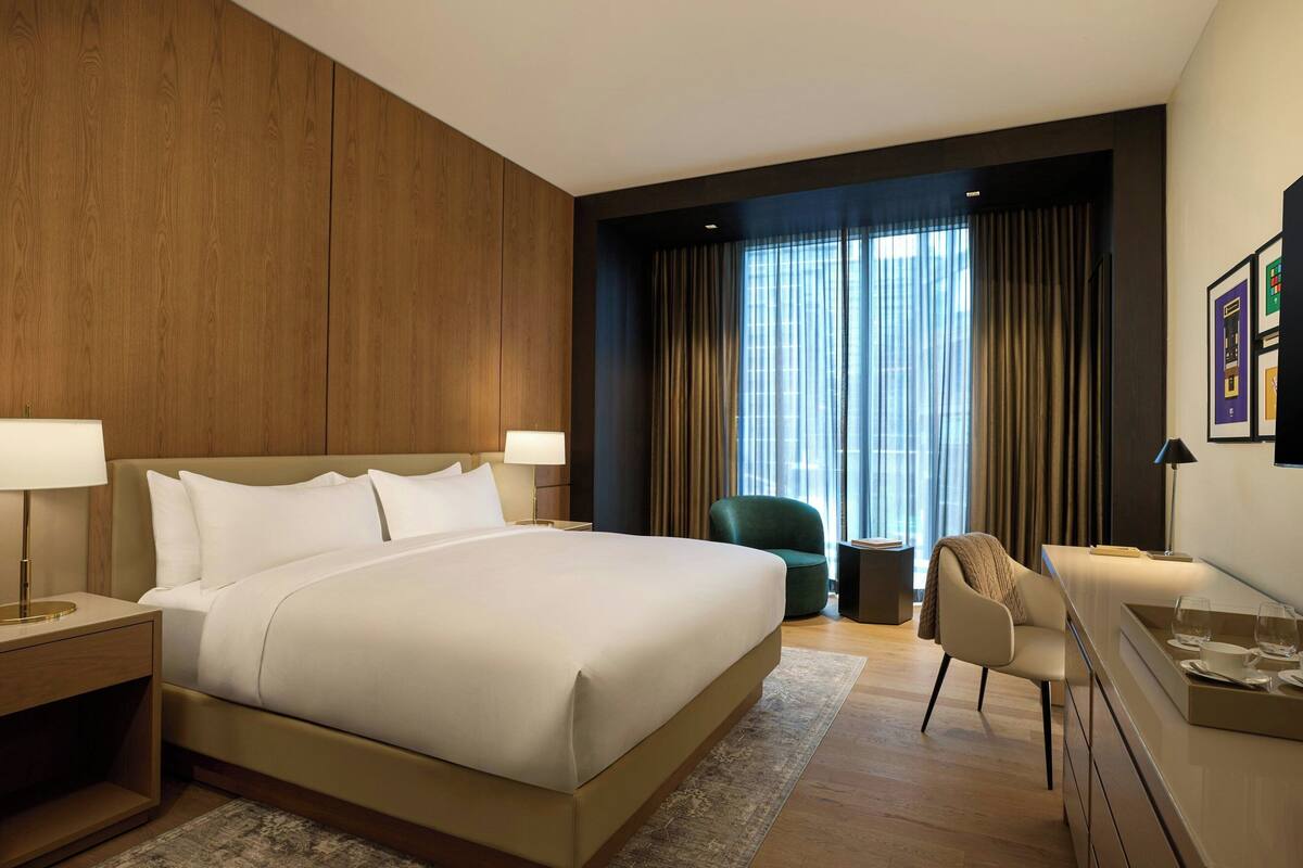 Habitación de hotel moderna con una cama grande, un escritorio, una silla y vistas a la ciudad a través de amplios ventanales.