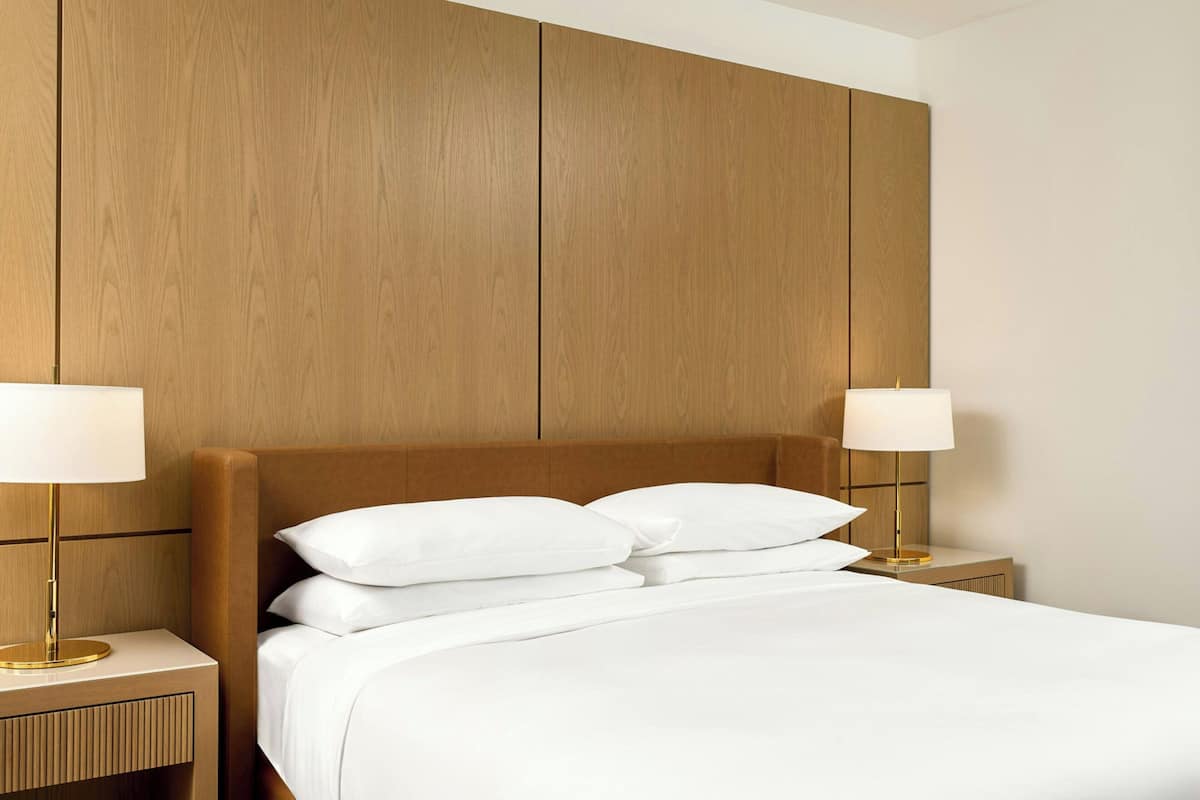Una cama bien hecha con sábanas blancas y dos lámparas de noche, frente a un cabecero y mesitas de madera.