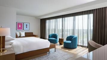 A modern hotel room with a large bed, two blue armchairs, a brown sofa, and a view of the outdoors through sheer curtains.