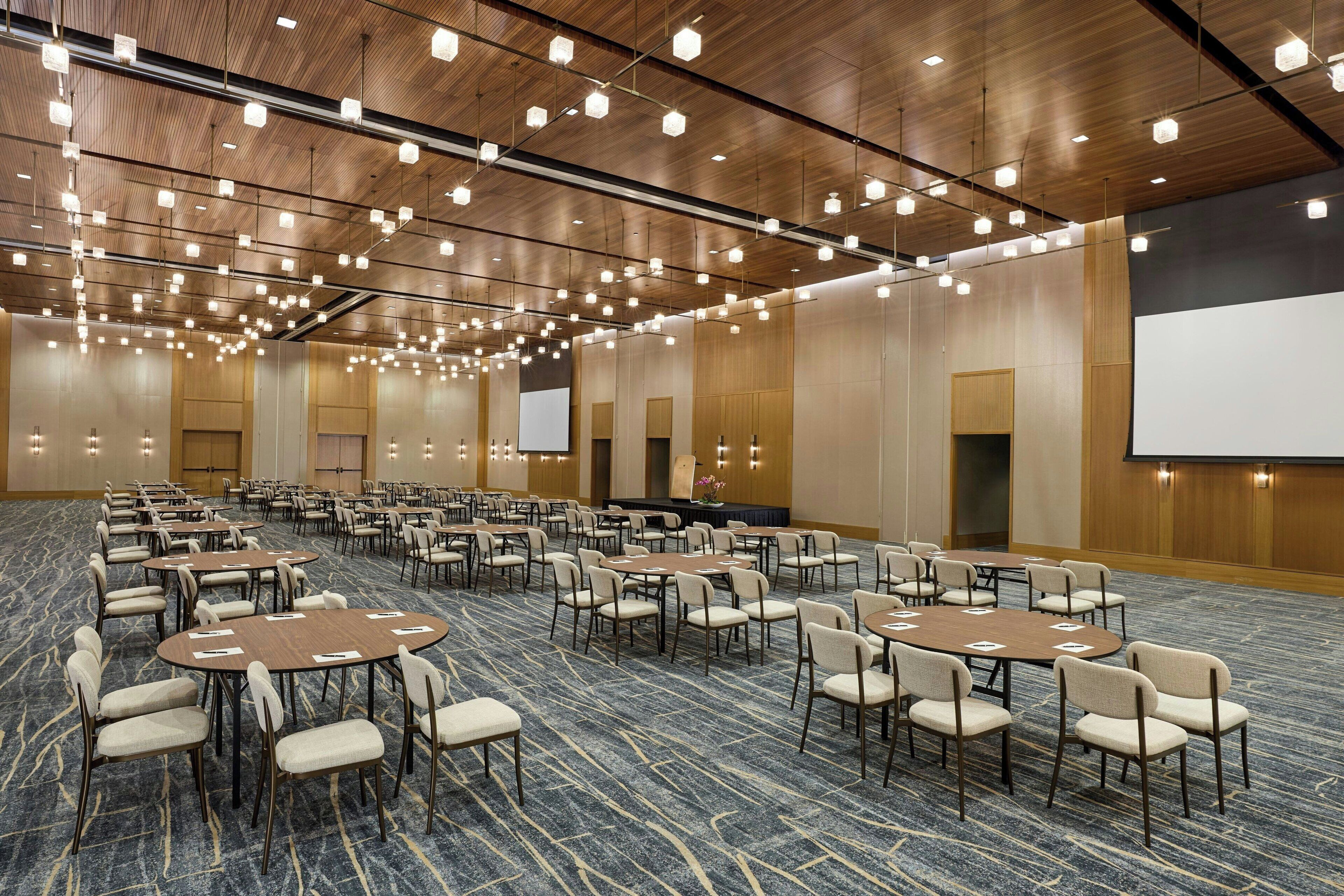 Une grande salle de conférence dotée de nombreuses tables et chaises, d’un plafond en bois avec des luminaires suspendus et d’un grand écran sur l’un des murs.
