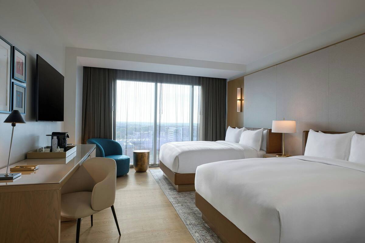 Habitación de hotel con dos camas, un escritorio, una silla, un televisor y un ventanal grande con cortinas.