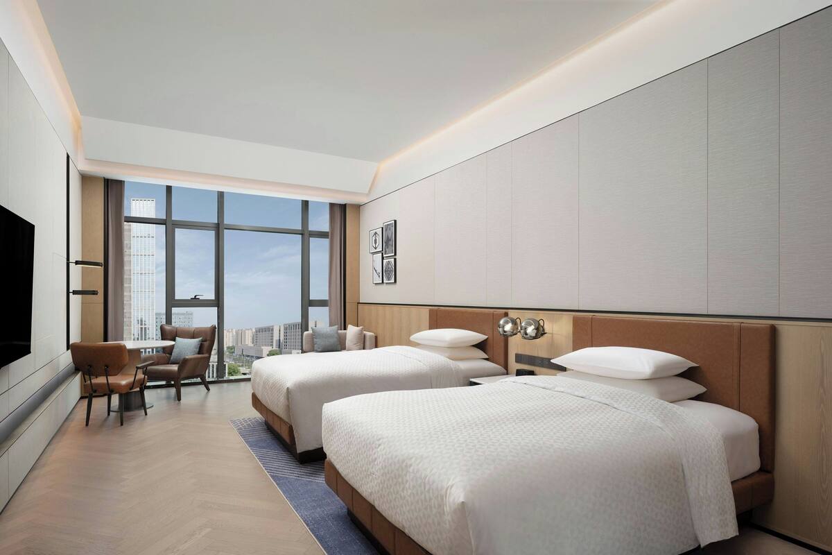 Een moderne hotelkamer met twee bedden, een eethoek en een groot raam met uitzicht op de stad.