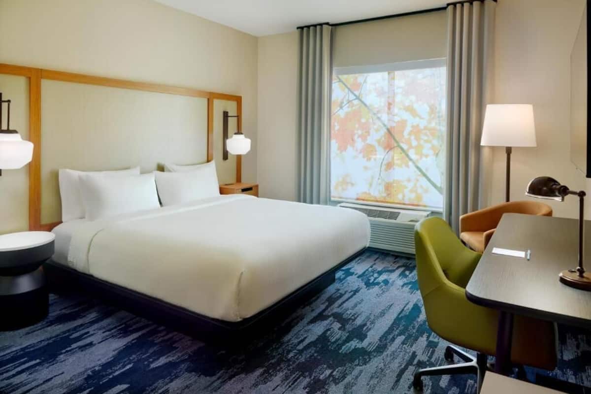 A hotel room with a large bed, a desk, a chair, a lamp, and a window with curtains.