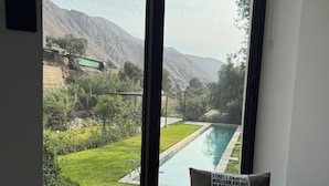 Habitación con un ventanal grande que da a una piscina y montañas. En primer plano, un banco de madera con un cojín.