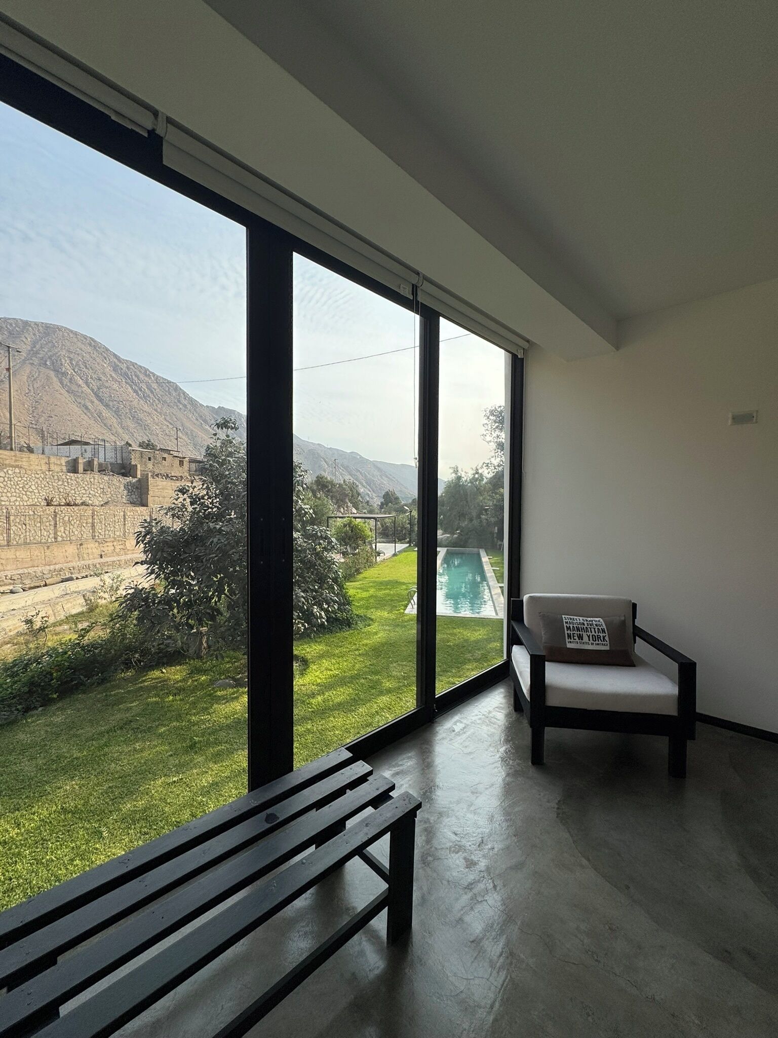 A modern room with large windows, a bench, a chair, and a view of a swimming pool and mountains.