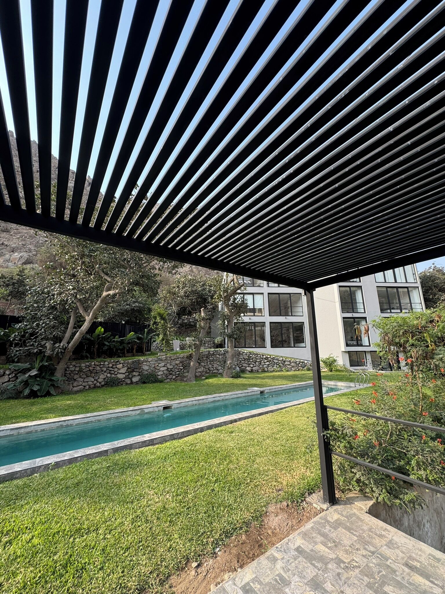A covered outdoor area with a pool, stone wall, and modern buildings in the background.