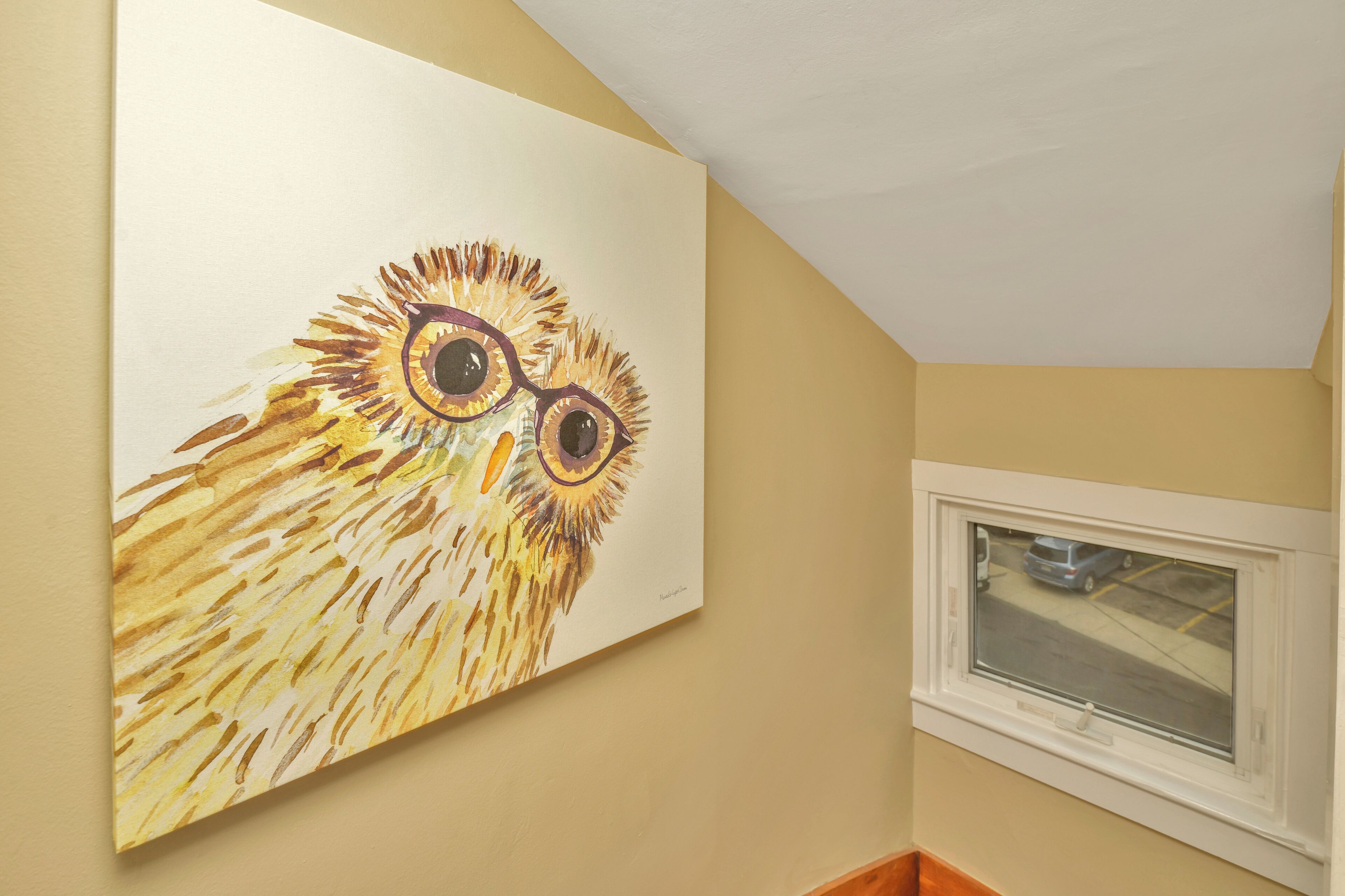 A room with a large painting of an owl on the wall, a window with a view of a street, and a wooden floor.