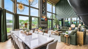 Un vaste espace de vie comprenant une grande table à manger, des sièges confortables, une cheminée et de grandes fenêtres donnant sur l’extérieur.