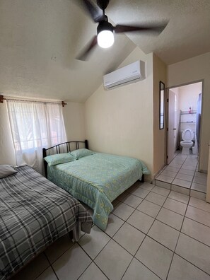 Une chambre avec deux lits, un ventilateur de plafond, un appareil de climatisation et une salle de bain visible à travers une porte ouverte.