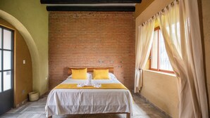 Una habitación de hotel con pared de ladrillo, cama con ropa blanca y amarilla, y ventana con cortinas.