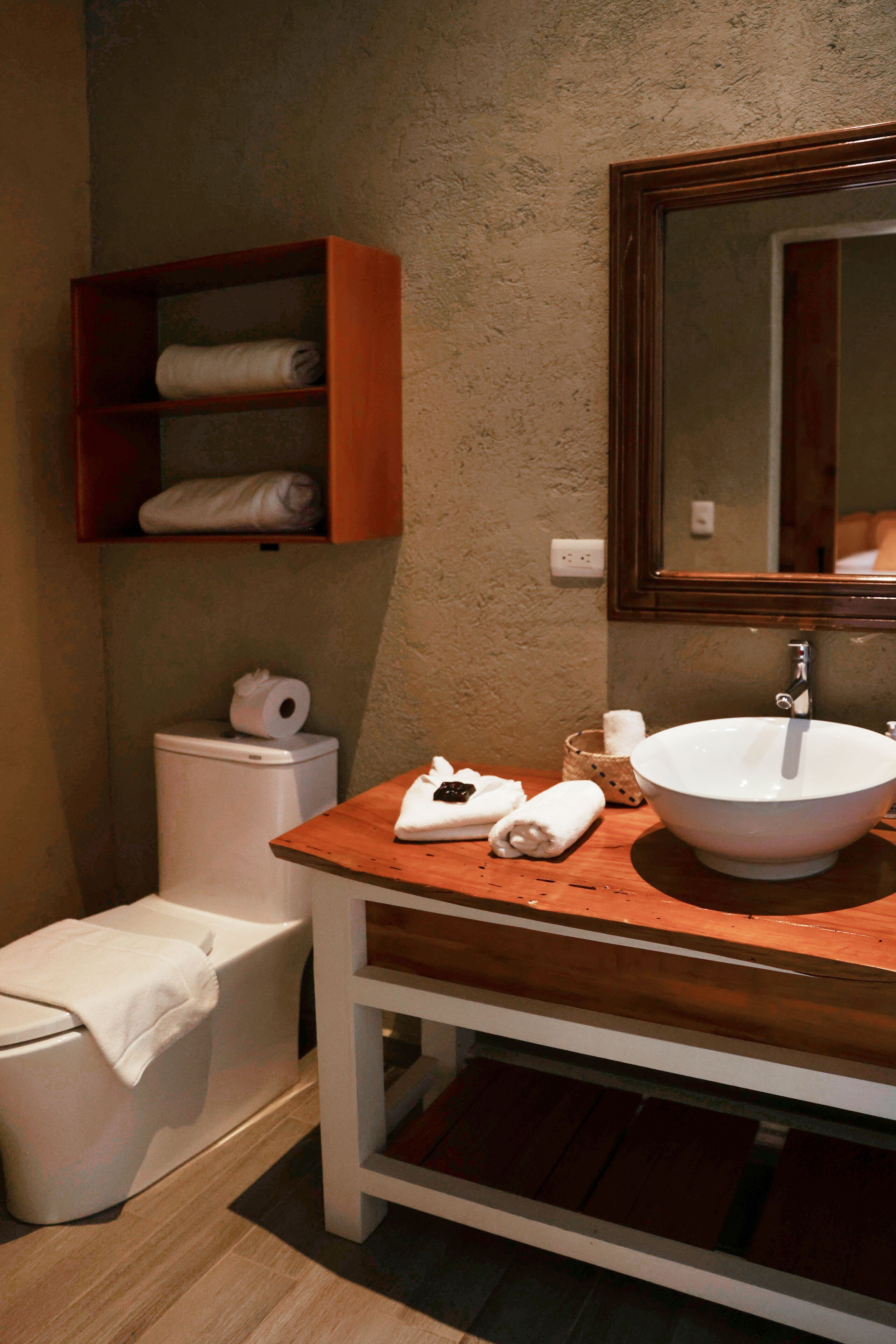 A bathroom with a wooden vanity, a white sink, a mirror, a toilet, and a shelf with towels.