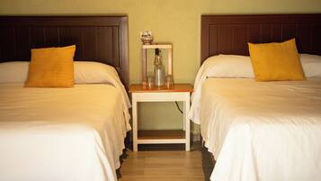 Habitación de hotel con dos camas, cada una con ropa blanca y una sola almohada amarilla. Una pequeña mesita de noche de madera con una botella de vidrio y un cuenco se encuentra entre las camas.