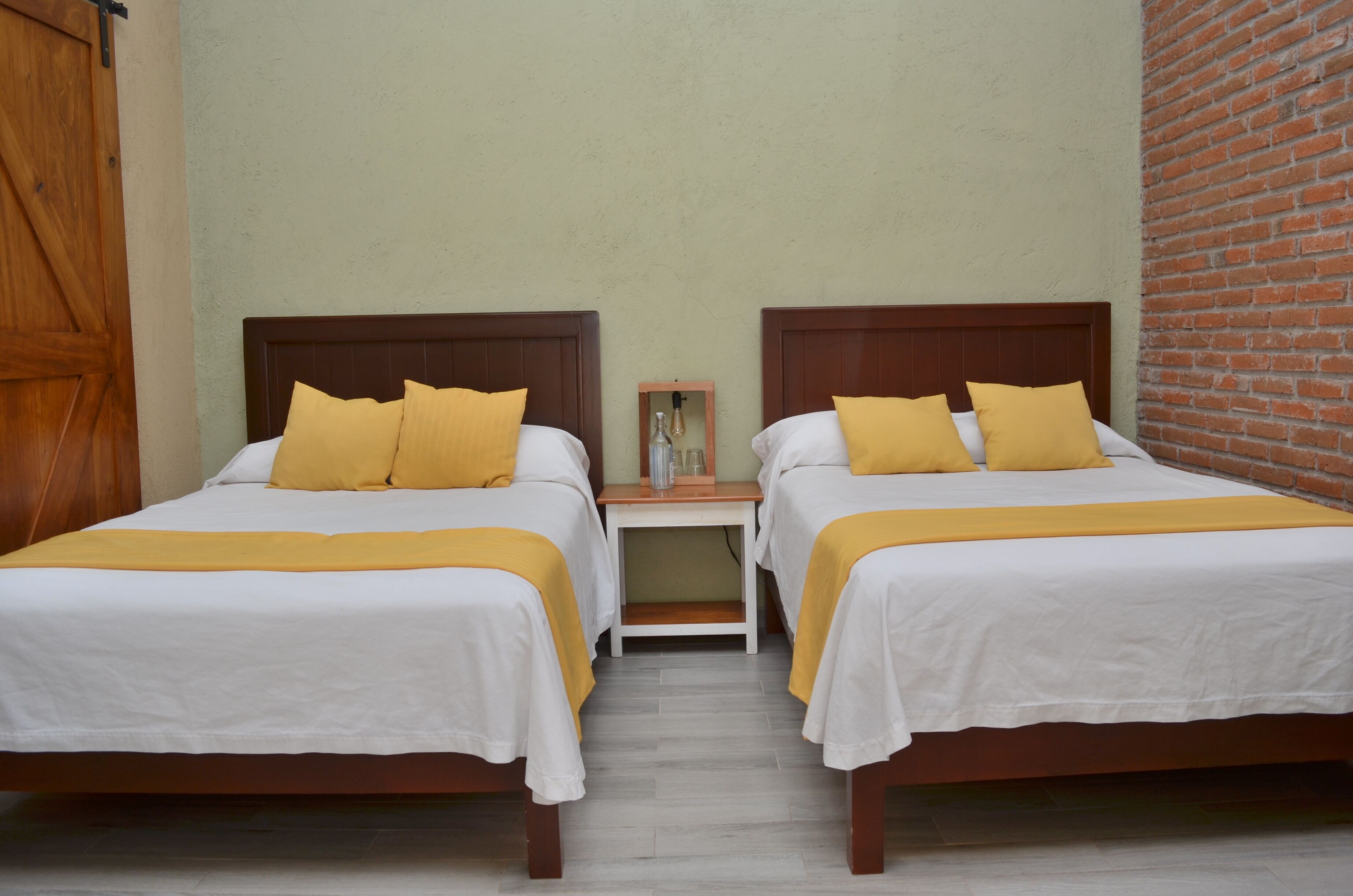 Habitación doble con dos camas, cada una con cabecero de madera, ropa de cama blanca y almohadas amarillas. Entre las camas hay una mesita de noche de madera con un adorno encima.