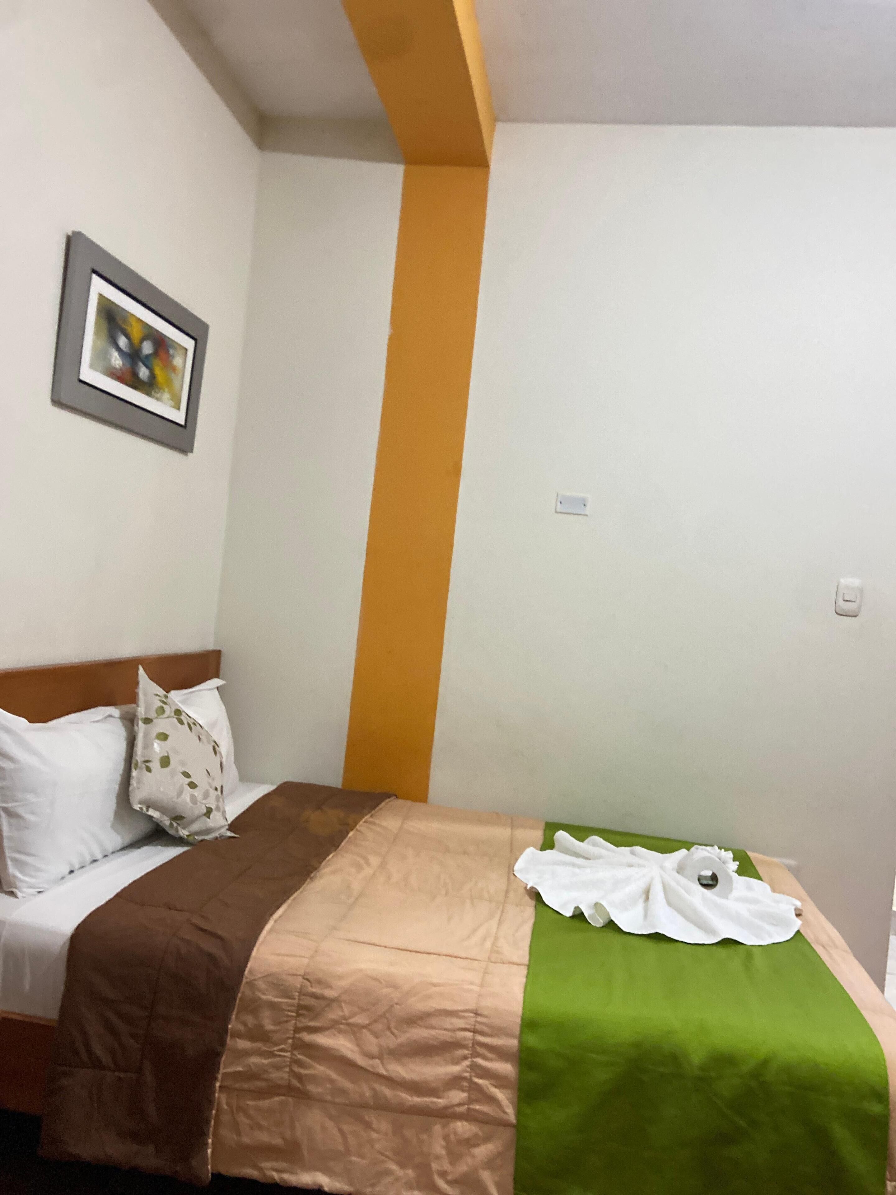 A hotel room with a bed, a green and beige bedspread, white pillows, and a framed picture on the wall.