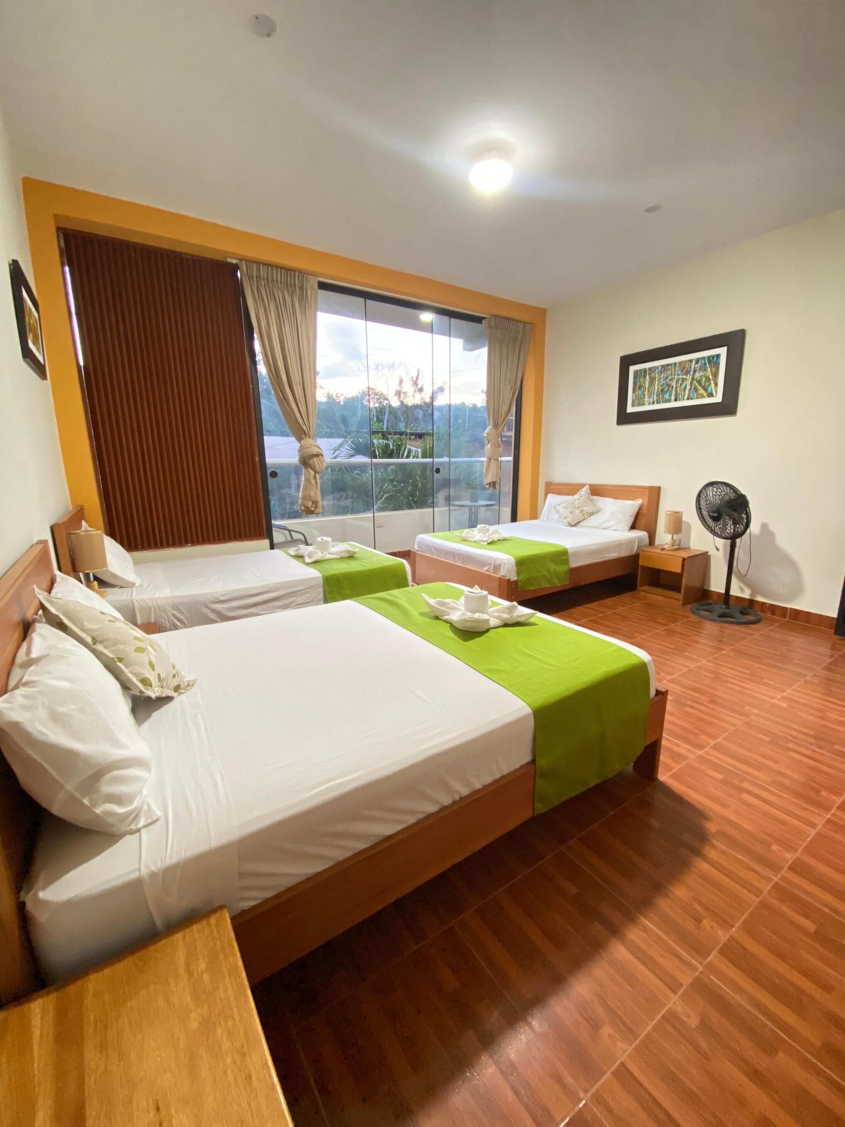 A hotel room with two beds, a wooden floor, a ceiling fan, and a window with curtains.