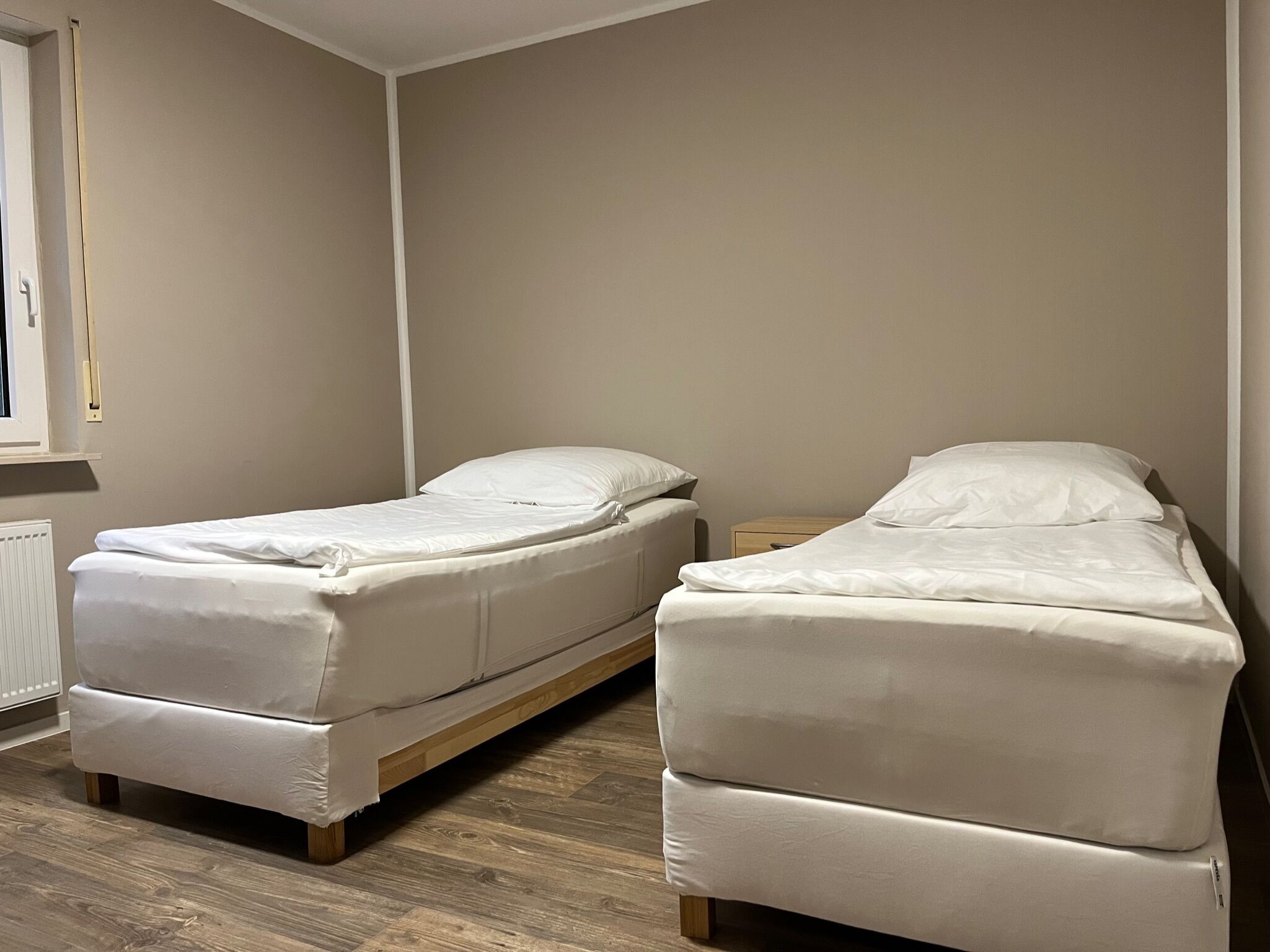 A room with two single beds, a wooden floor, a radiator, and a window with a curtain.