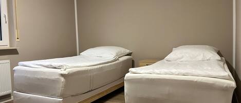 A room with two single beds, a wooden floor, a radiator, and a window with a curtain.