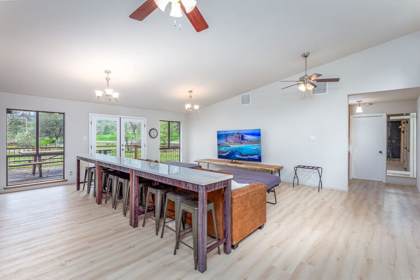 Una habitación amplia con una larga mesa de comedor, taburetes de bar, un ventilador de techo y un gran ventanal con vistas al exterior.
