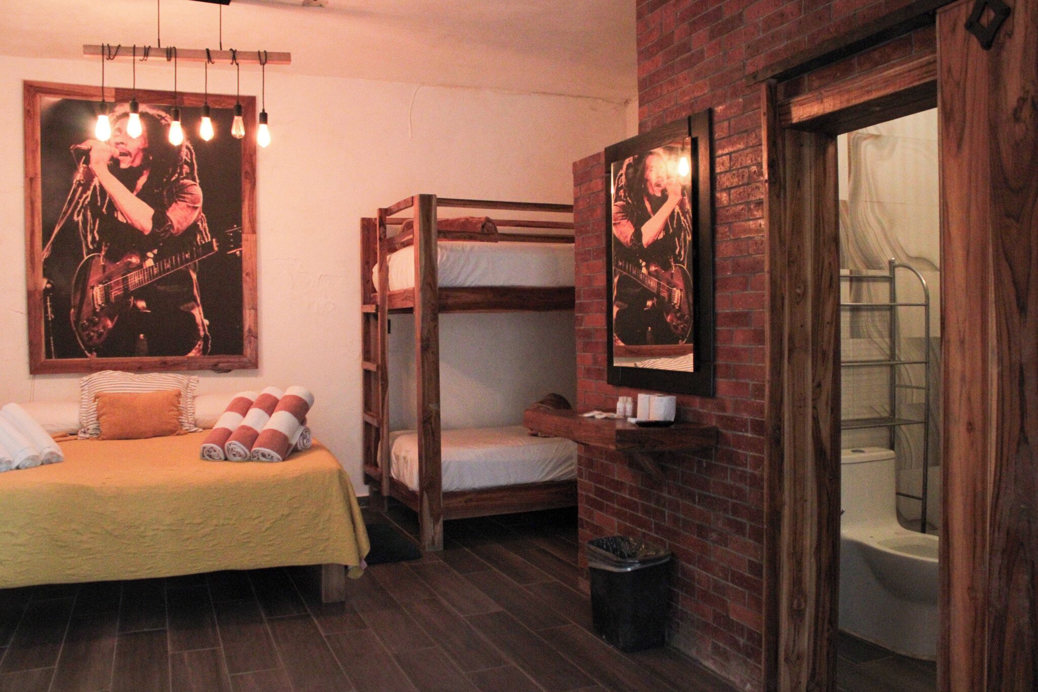 A room with a bunk bed, a single bed with a yellow bedspread, and a brick wall. There are posters on the wall and a window with a view of the outside.
