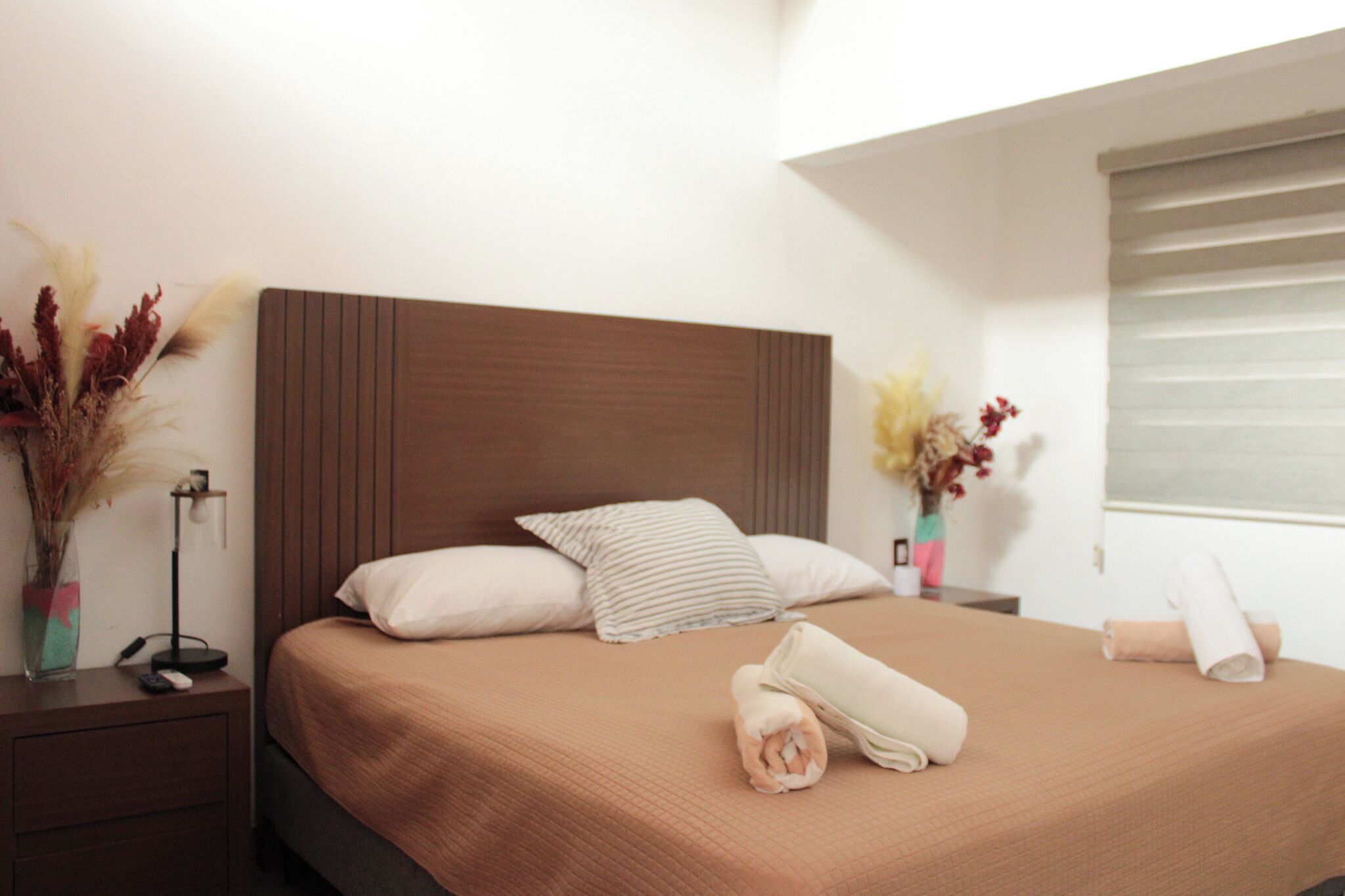 Una cama bien hecha con cabecera marrón, almohadas blancas y a rayas, y toallas enrolladas en una habitación moderna y limpia con ventana y jarrones decorativos.