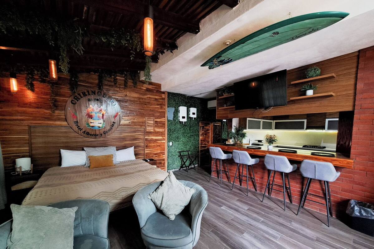 Quarto com cama, duas cadeiras, espaço bar com banquetes, uma prancha de surf e televisão.