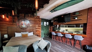 A room with a bed, two chairs, a bar area with stools, a surfboard, and a television. - Hotel Chiringuito de Palo (Aticama)