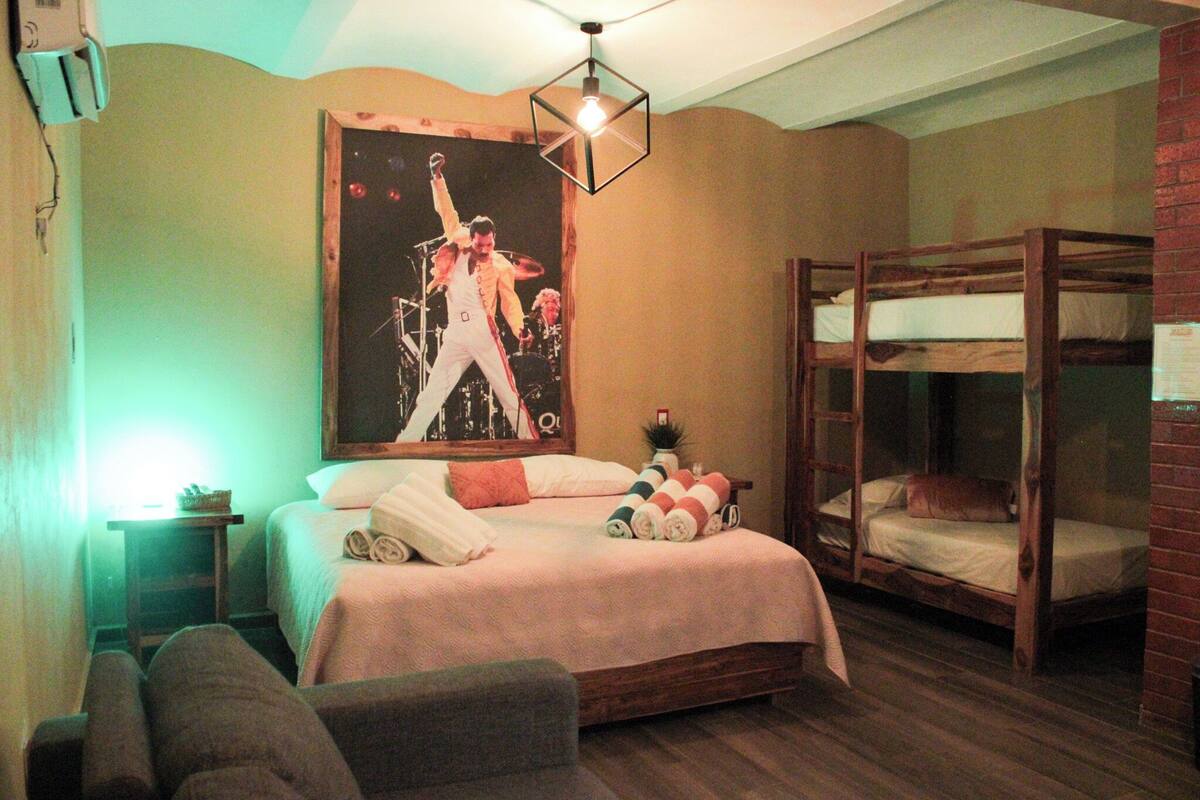 Um quarto com uma cama de solteiro, uma cama individual com toalhas enroladas, um cartaz na parede e uma pequena mesa com uma planta.