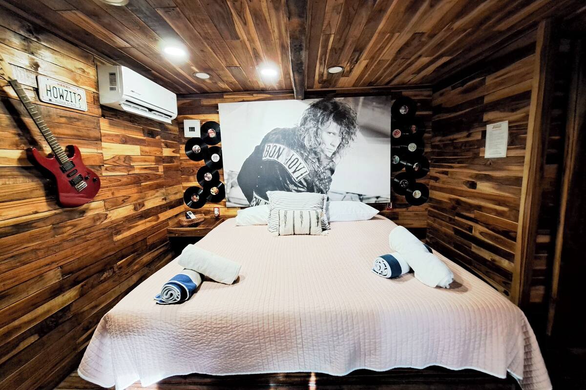 Quarto com paredes de madeira, cama com colcha acolchoada, uma guitarra encostada na parede e ar condicionado fixado na parede.