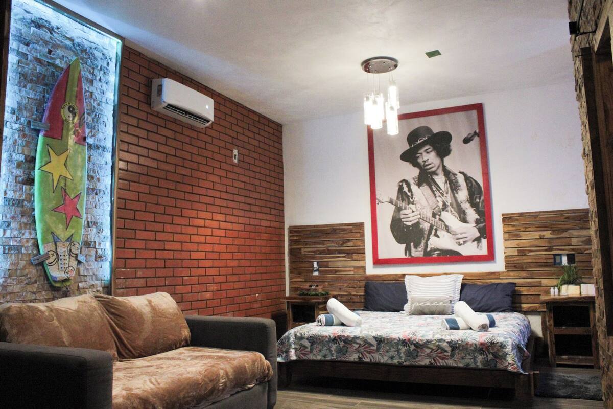 Quarto com parede de tijolos, cama com colcha florida, cabeceira de madeira, mesa de cabeceira, uma cadeira e ar-condicionado fixado na parede.