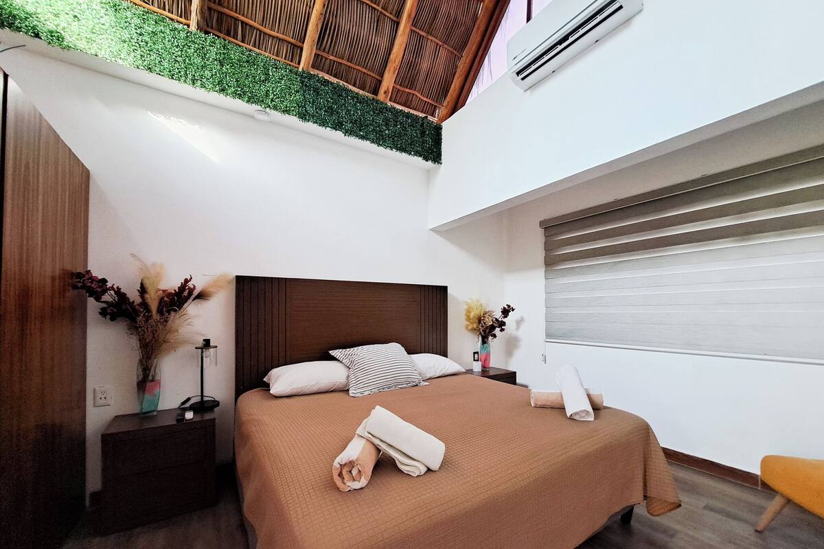 Quarto moderno com cama grande, mesa de cabeceira, criadeira com abajur, janela com persianas e um pequeno banco.