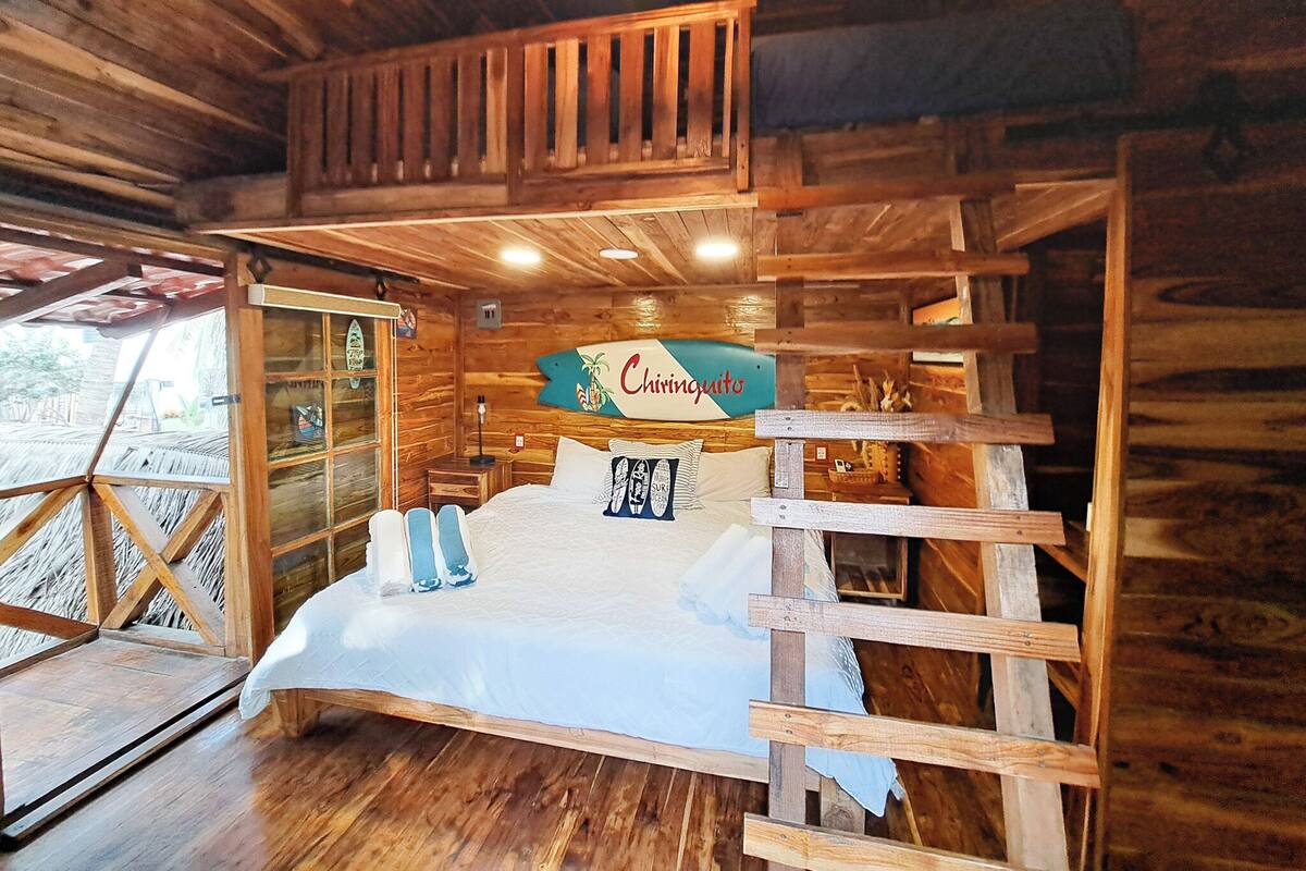 Quarto estilo cabana de madeira, com uma cama, um letreiro indicando "Chiringuito" e uma escada de madeira.