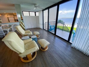 A spacious balcony with a view of the ocean, furnished with beige leather chairs and round wooden tables.