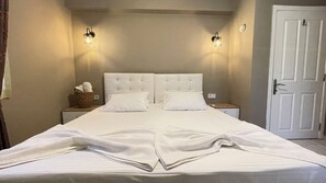 Ein Doppelbett mit weißem Kopfteil und Fußteil, zwei Nachttische mit Lampen, ein Korb mit Handtüchern und eine weiße Tür.