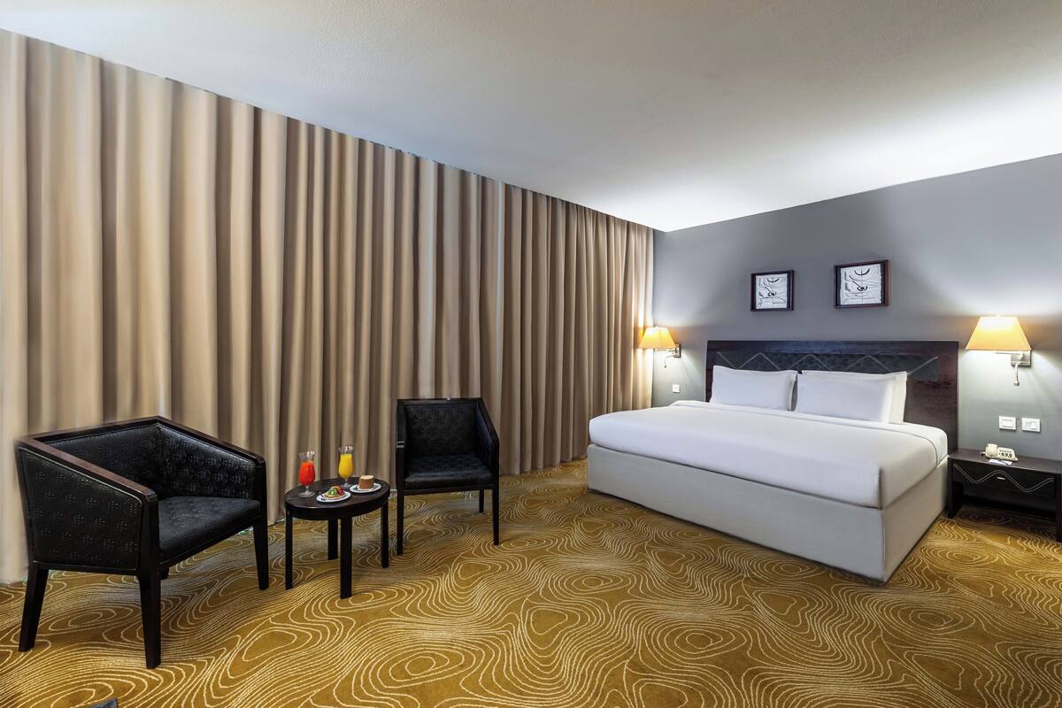 Habitación de hotel con una cama grande, dos sillas, una mesita con bebidas y una alfombra estampada.