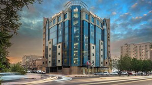 A modern building with a glass facade and multiple floors, surrounded by trees and parked cars. - Razana Al Jubail (Al Jubail)