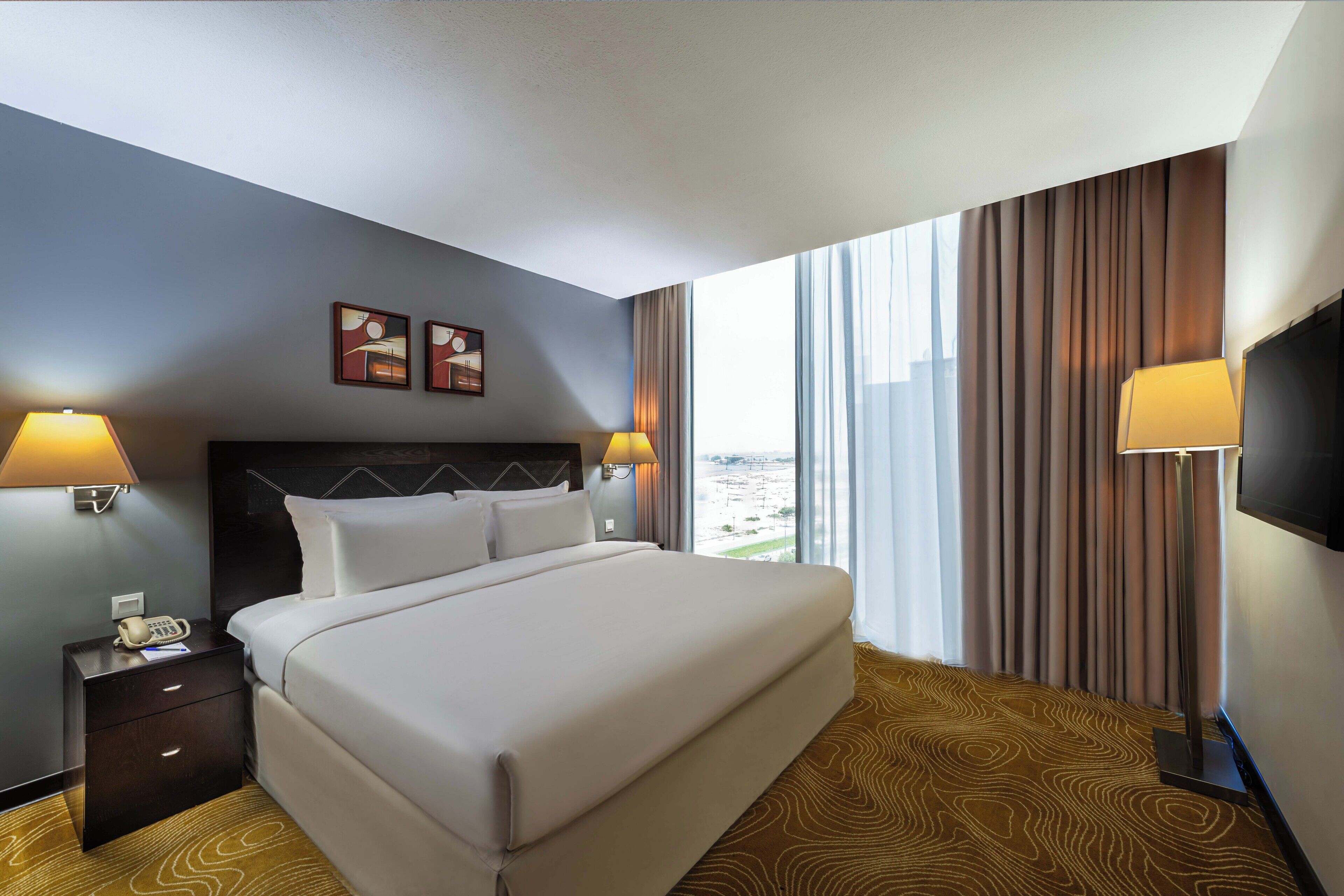 Habitación de hotel con una cama grande, dos lámparas de noche, una mesita de noche con teléfono y vistas a la ciudad a través de amplios ventanales.