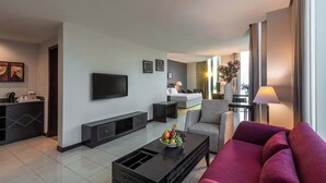 A modern living room with a flat-screen TV, a sofa, a coffee table with fruit, and a dining area with a lamp. - Razana Al Jubail (Al Jubail)