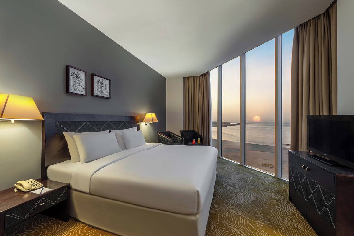 Habitación de hotel con una cama grande, dos lámparas de noche, un televisor sobre un mueble y vistas a la puesta de sol a través de grandes ventanales.