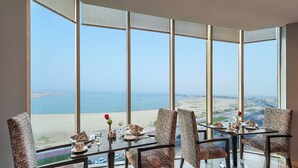 A dining area with a table set for four, overlooking a beach and cityscape. - Razana Al Jubail (Al Jubail)