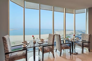 Comedor con mesa lista para cuatro personas, con vista a la playa y al paisaje urbano.